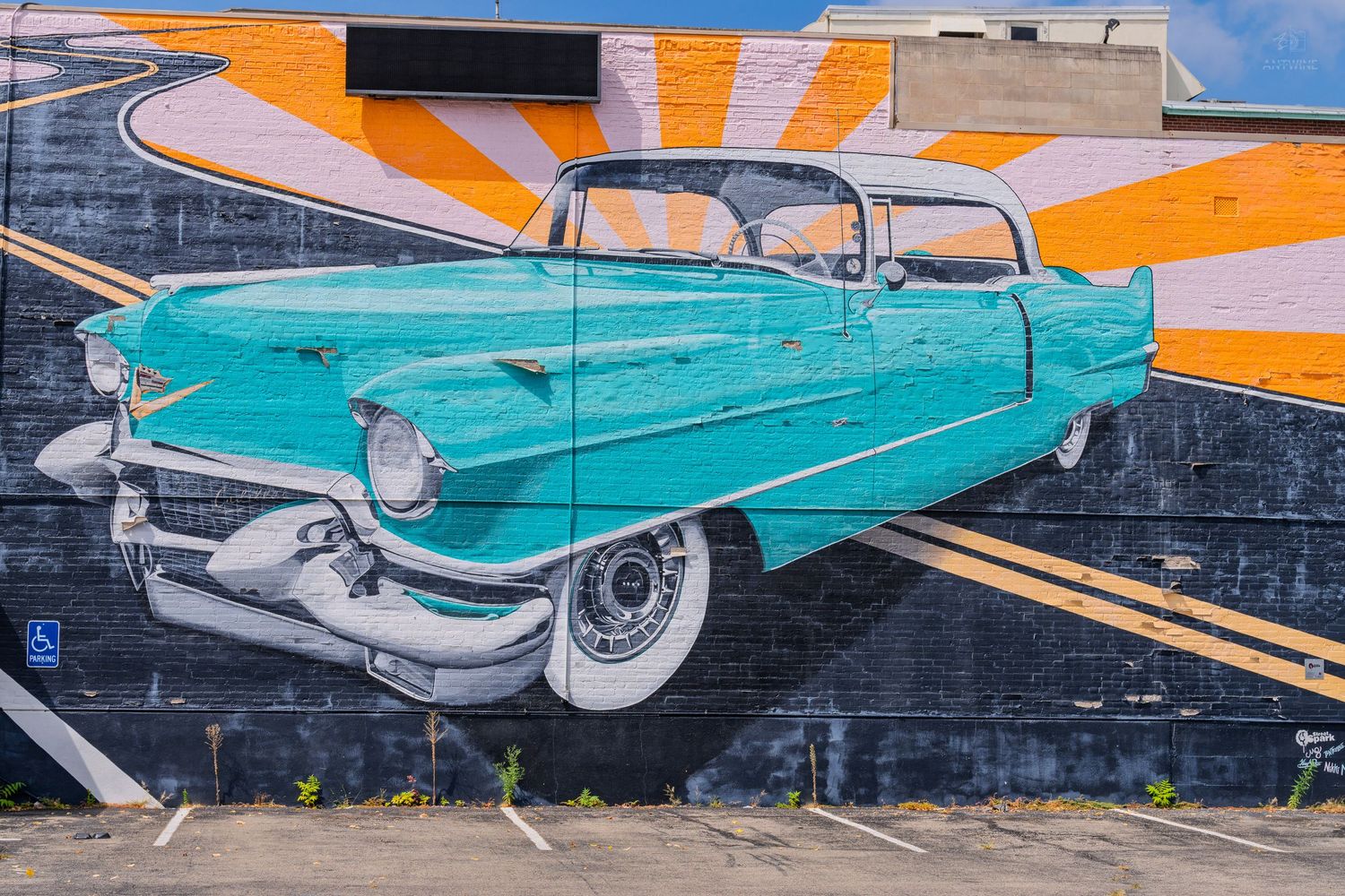 Vibrant street mural of a classic teal 1950s Cadillac convertible on a brick wall with orange sunburst background.