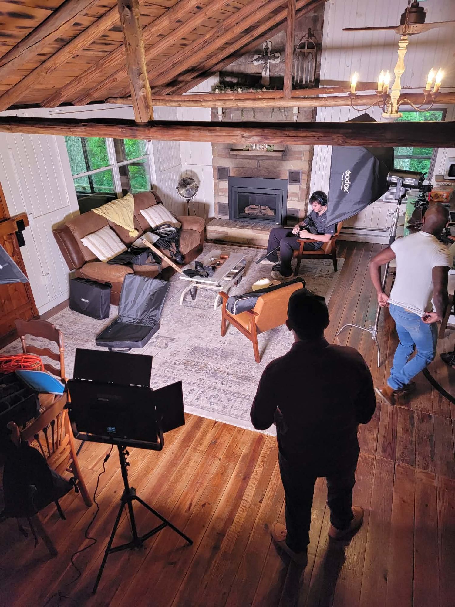 Film crew setting up a rustic cabin interior shoot with lighting equipment, fireplace, and hardwood floors.