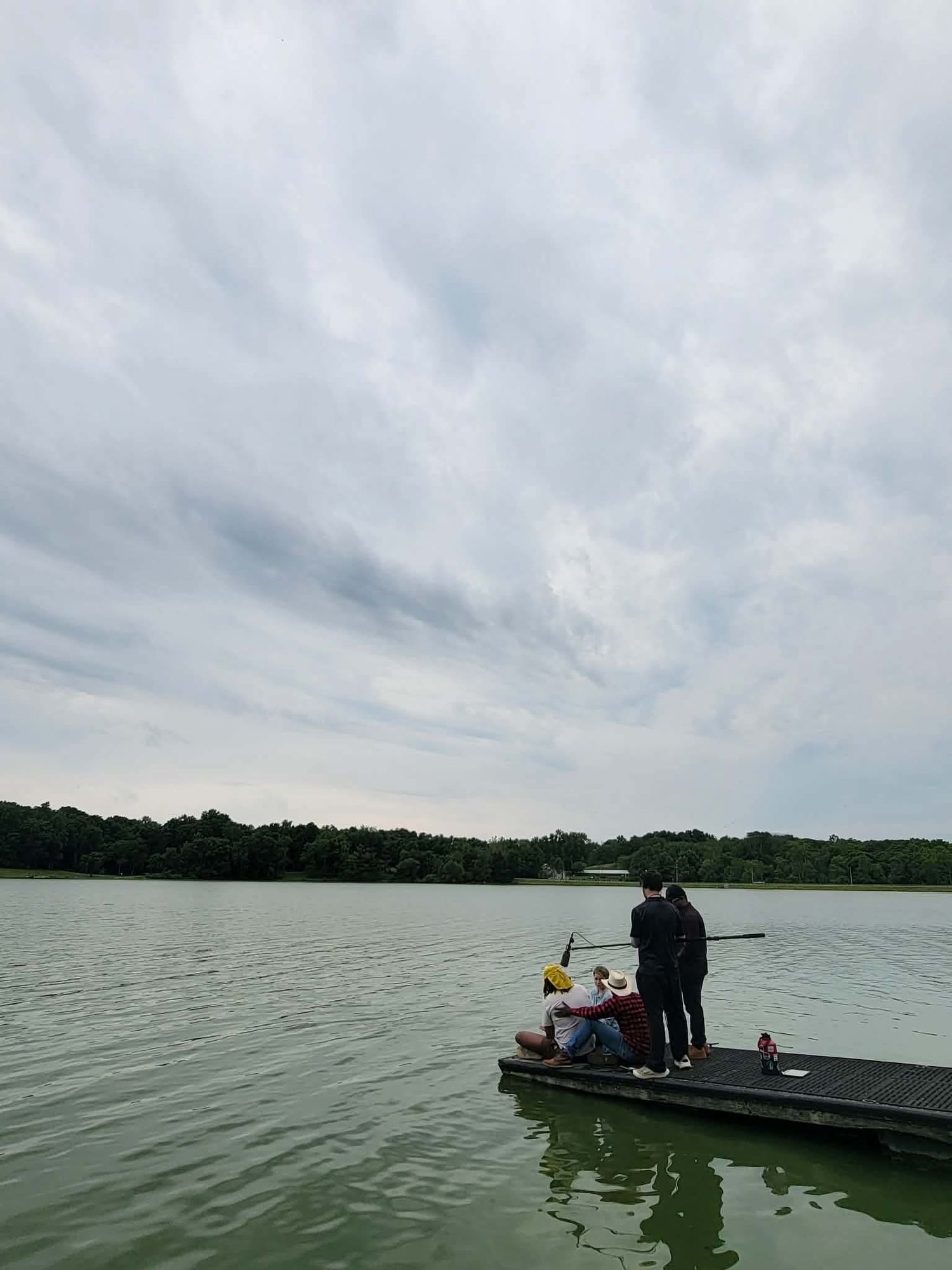 People fishing on a dock over a calm green lake under a cloudy sky, surrounded by lush trees.