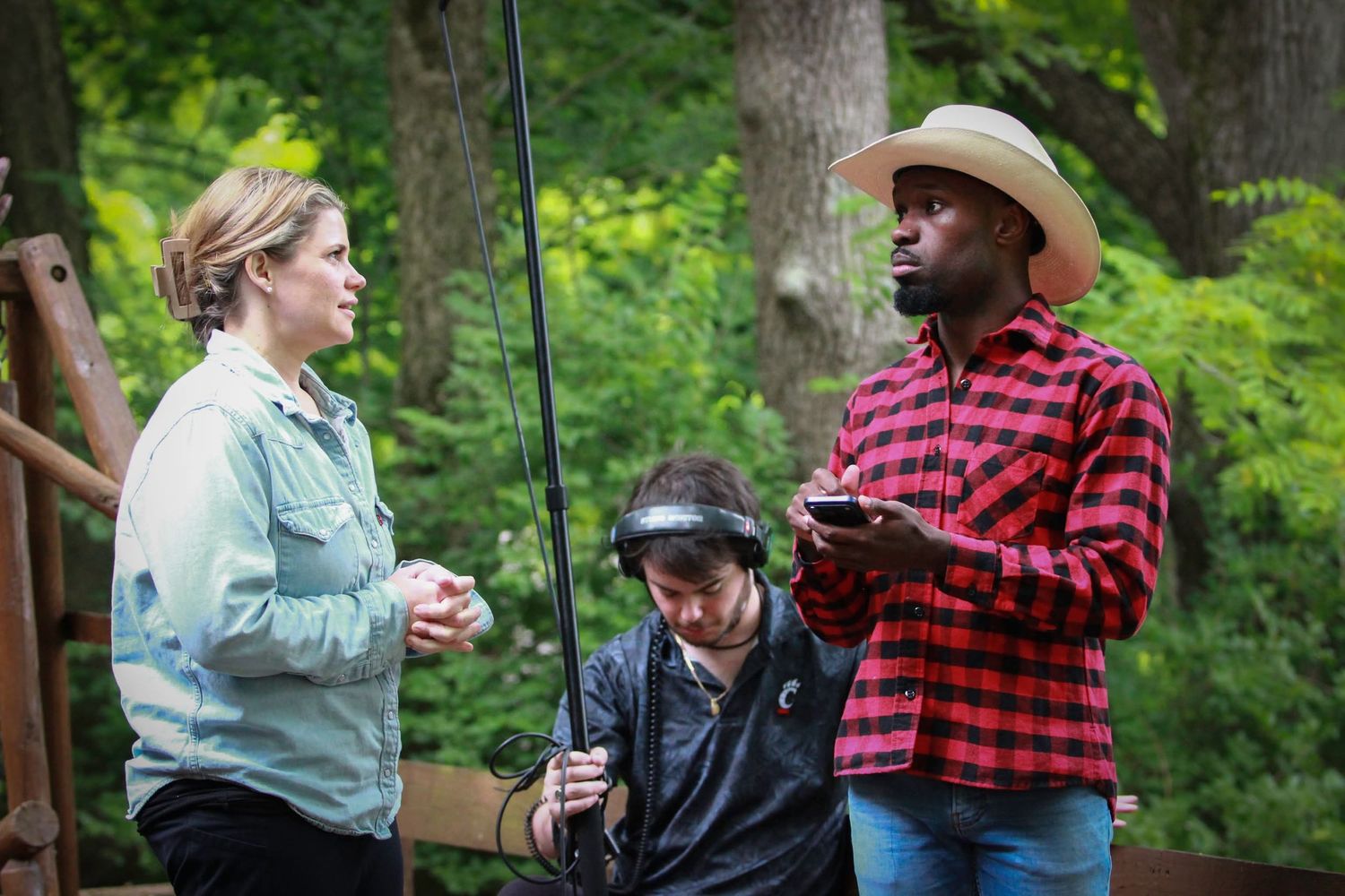 Three people collaborate outdoors in wooded area, one holding phone, during a film production.