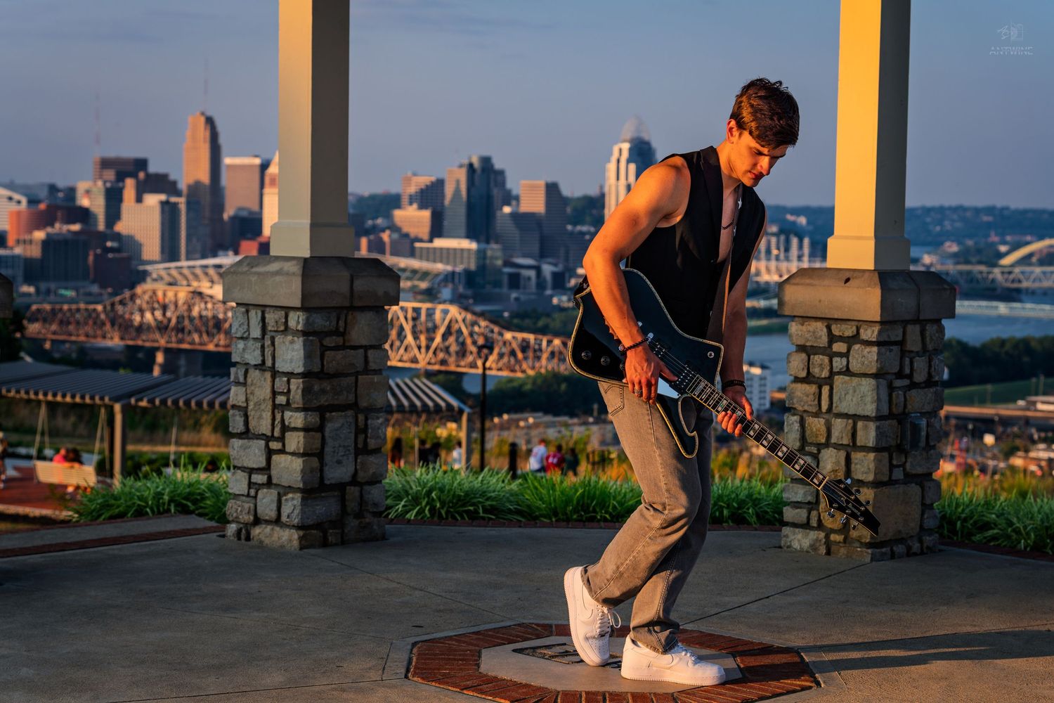 Young man playing electric guitar outdoors at golden hour with city skyline behind him.
