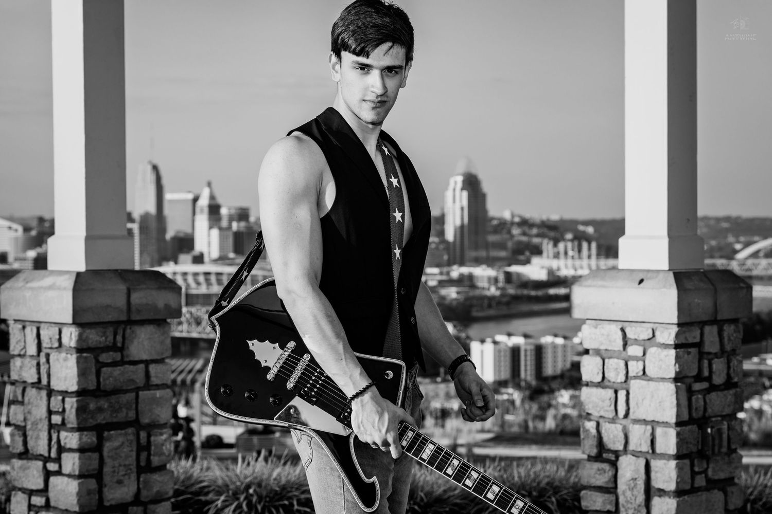 Young male musician holding electric guitar outdoors with city skyline backdrop in black and white.