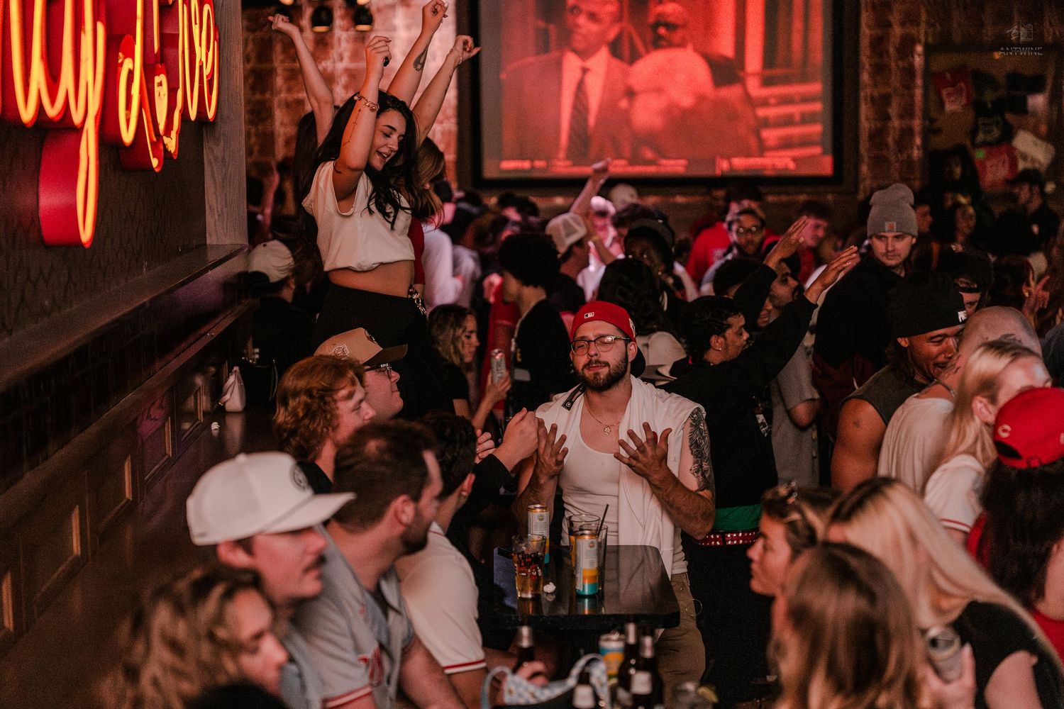Lively bar crowd socializes under neon lights with a large screen projection in the background.