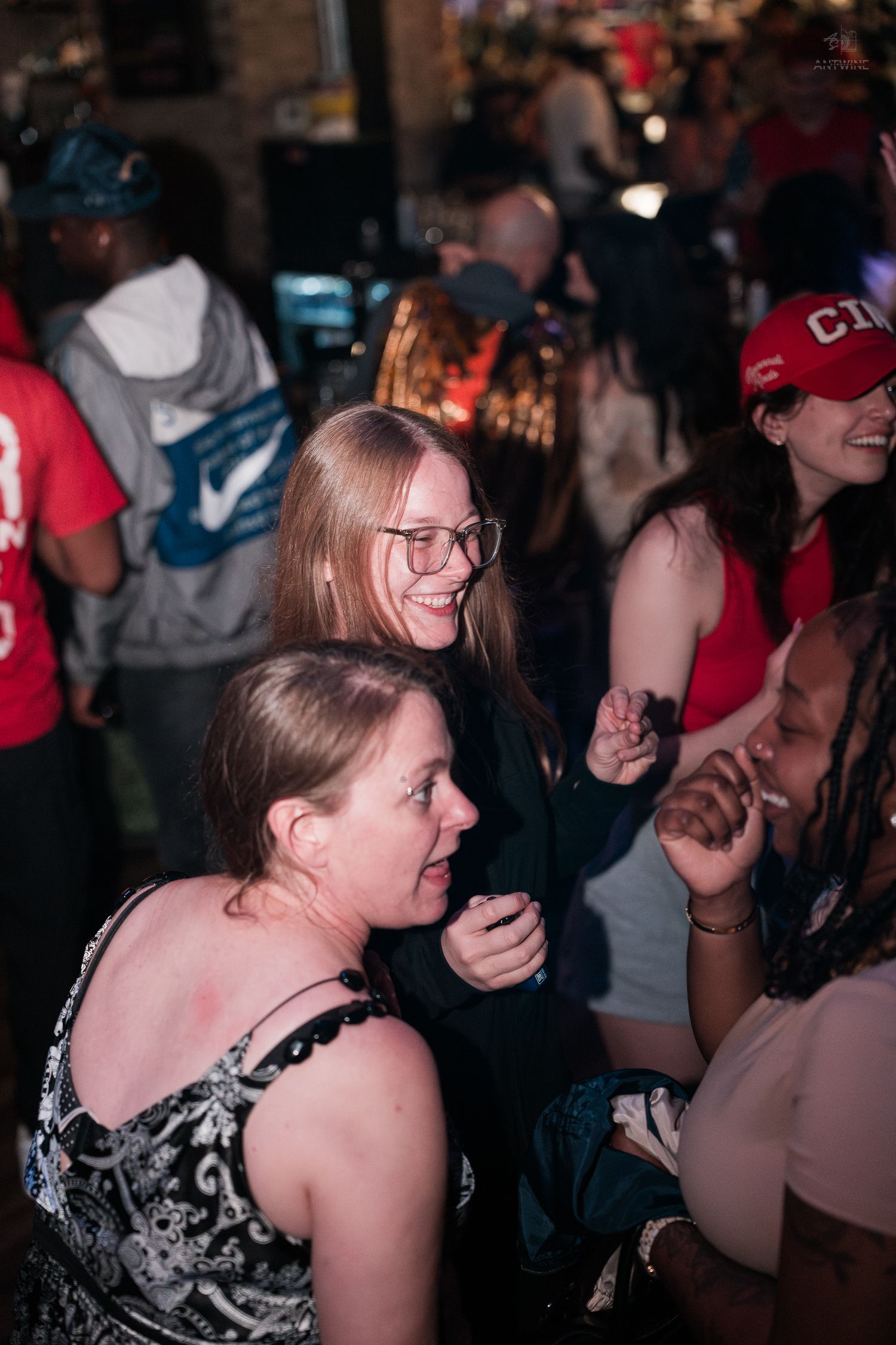 Women laughing and dancing together at a crowded indoor bar or nightclub party.