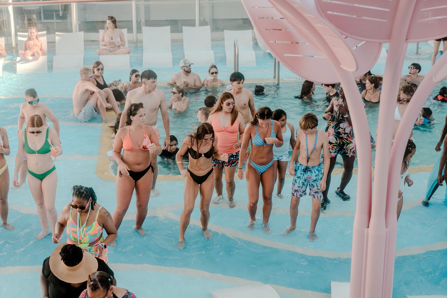 Cruise ship guests in colorful swimwear enjoying a pool deck dance party in shallow water.