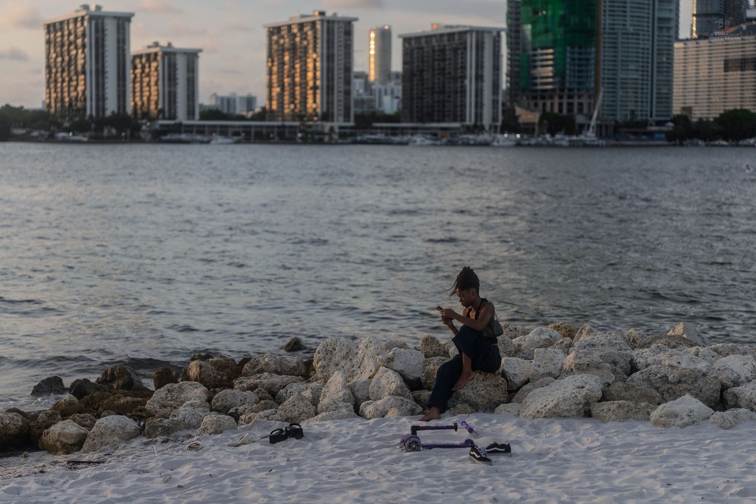 Woman sitting on rocky shoreline using phone next to an electric scooter with city skyline at dusk.