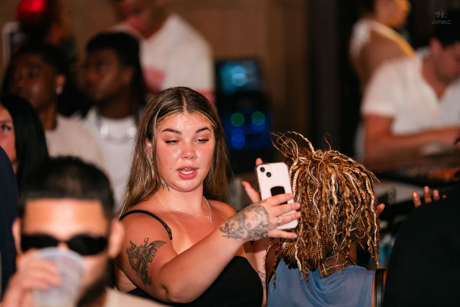 Woman with tattoos showing a phone to a person with dreadlocks at a party.
