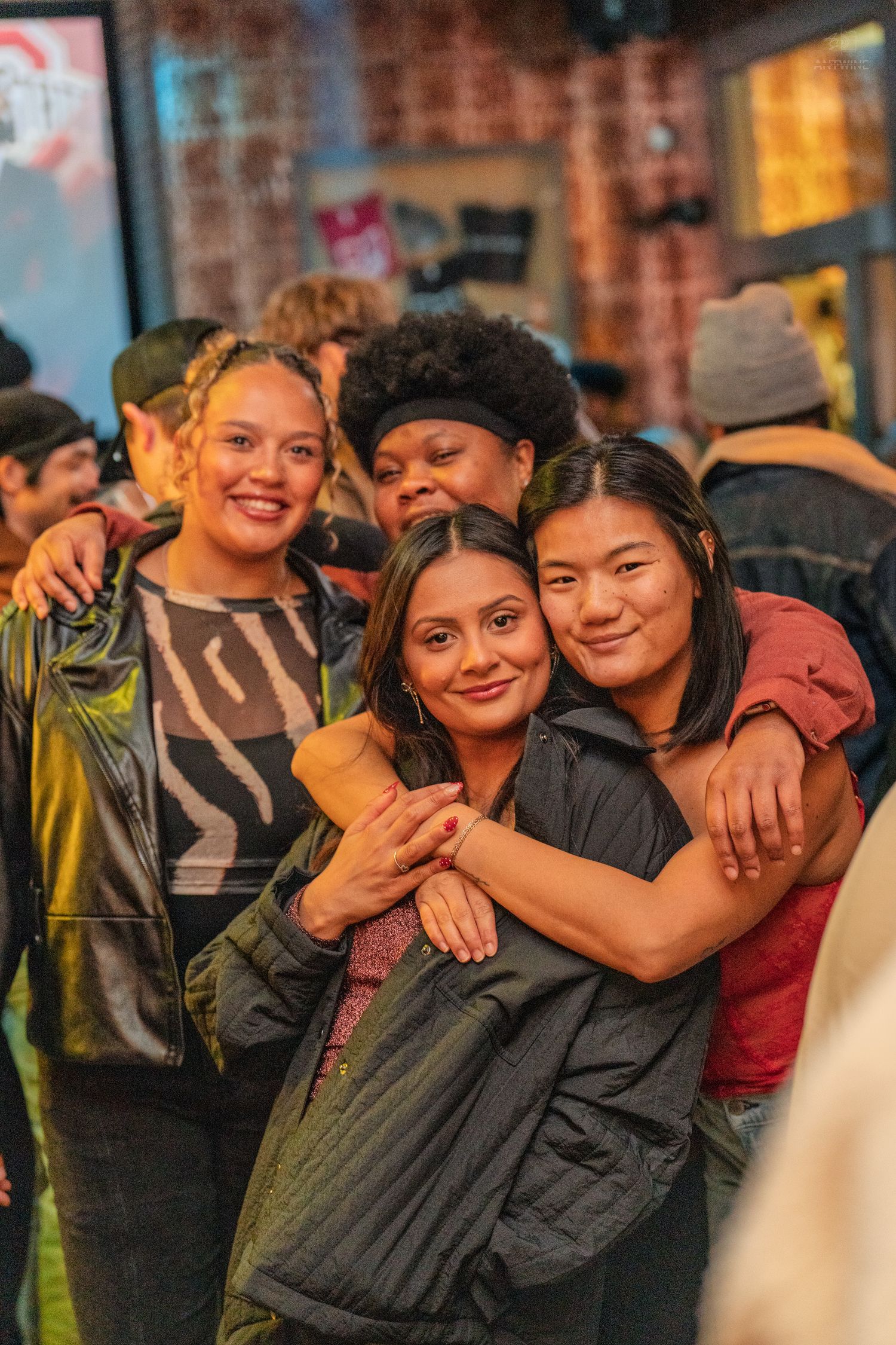 Four diverse young women smile and hug together outdoors at a nighttime social event.