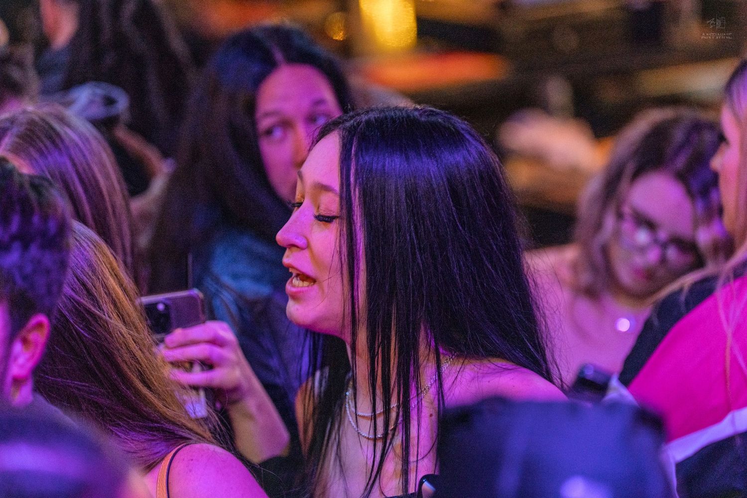 A woman with long dark hair looks at her phone under purple lights in a crowded venue.