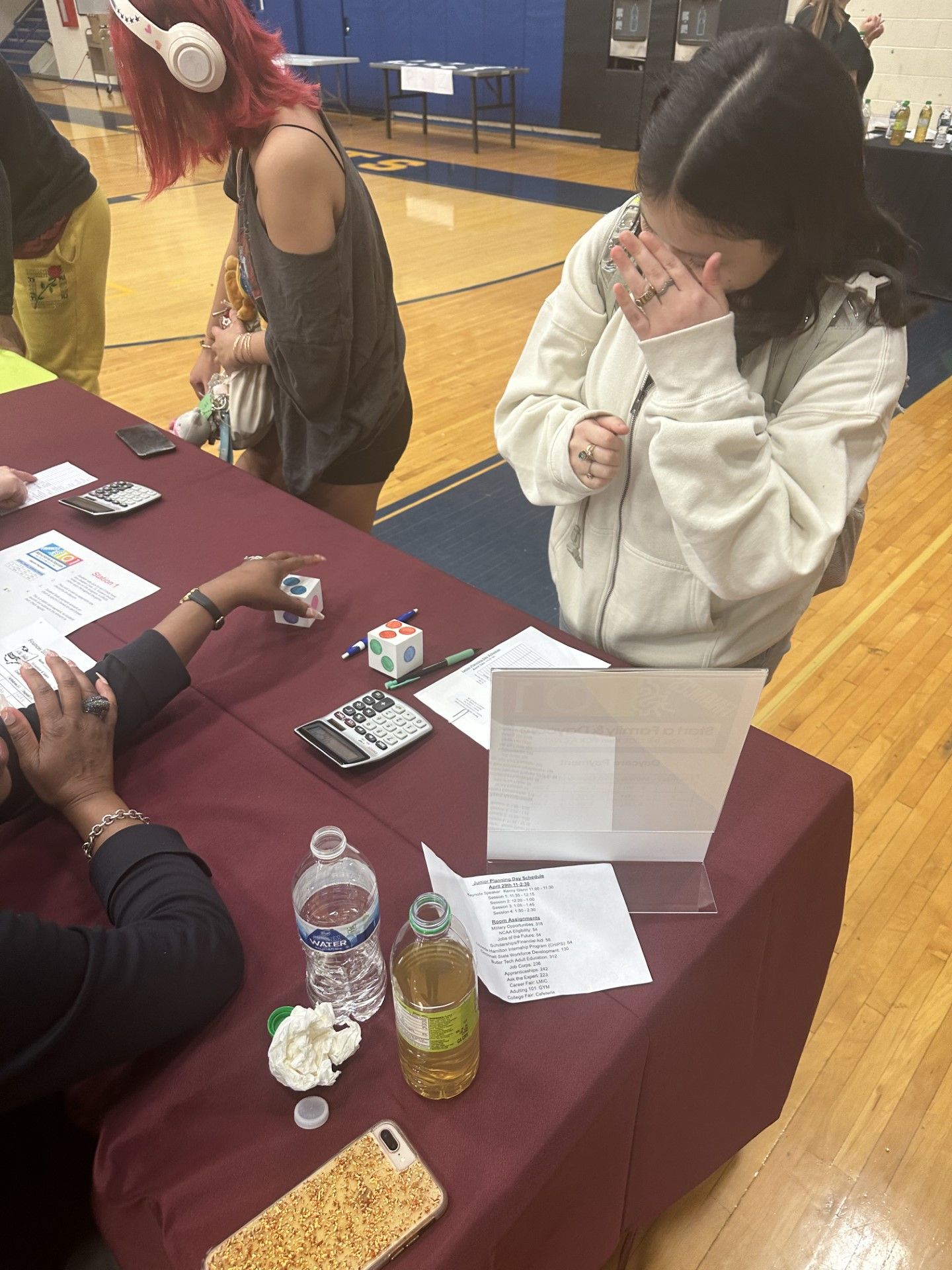 Students sit at a maroon-draped table in a school gym, reviewing papers and items during an educational fair event.