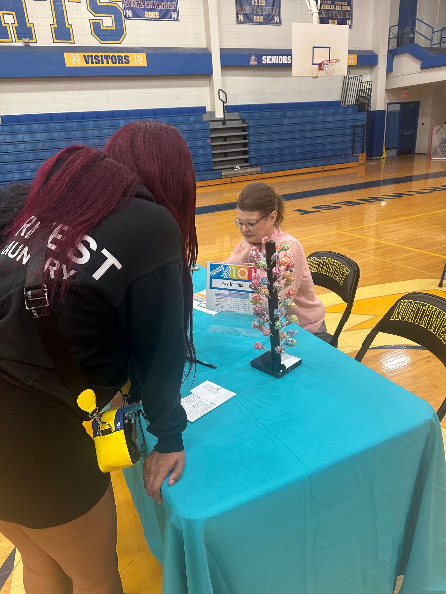 Two women at a teal table in a school gymnasium during a career fair event with a colorful display stand.