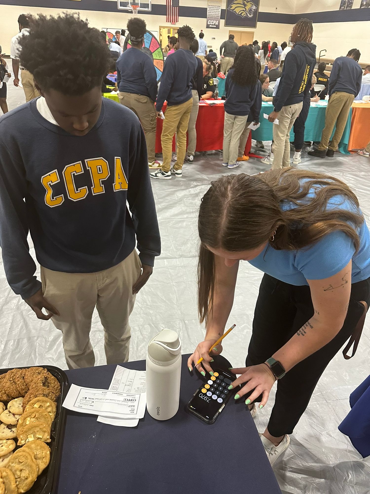 Students at a school fair event, one wearing ECPA sweatshirt, while a girl demonstrates a color test kit at a table.