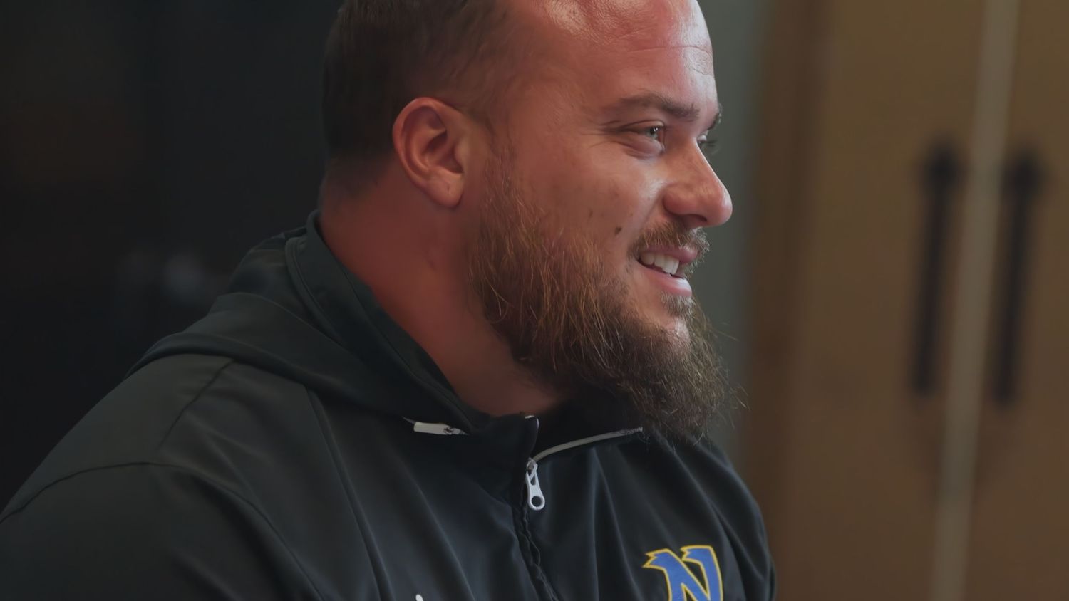 Smiling bearded man in a navy blue sports jacket with a logo, appearing relaxed and confident indoors.