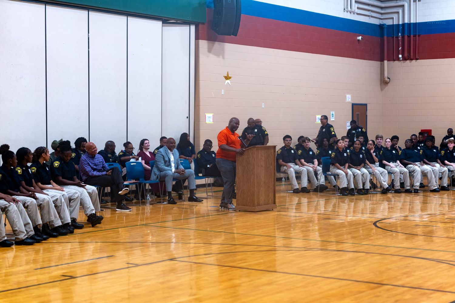 Students seated in gymnasium rows listen to a speaker at a podium during a school assembly event.