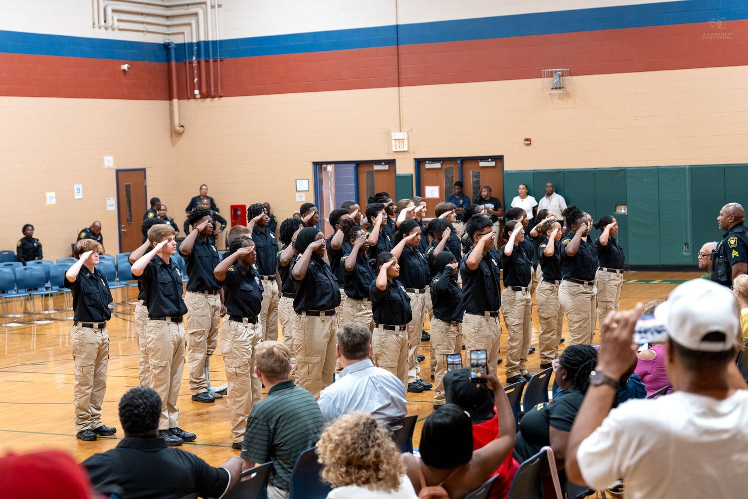 Group of uniformed cadets saluting during an indoor graduation ceremony in a gymnasium with seated audience.