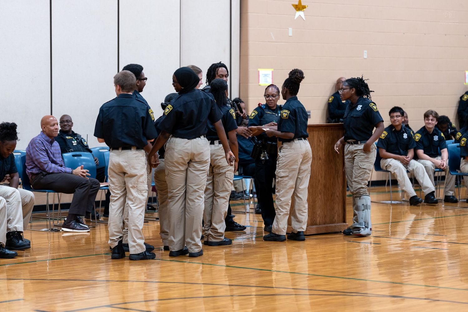 Students in uniform gather around a podium in a school gymnasium during an indoor assembly event.