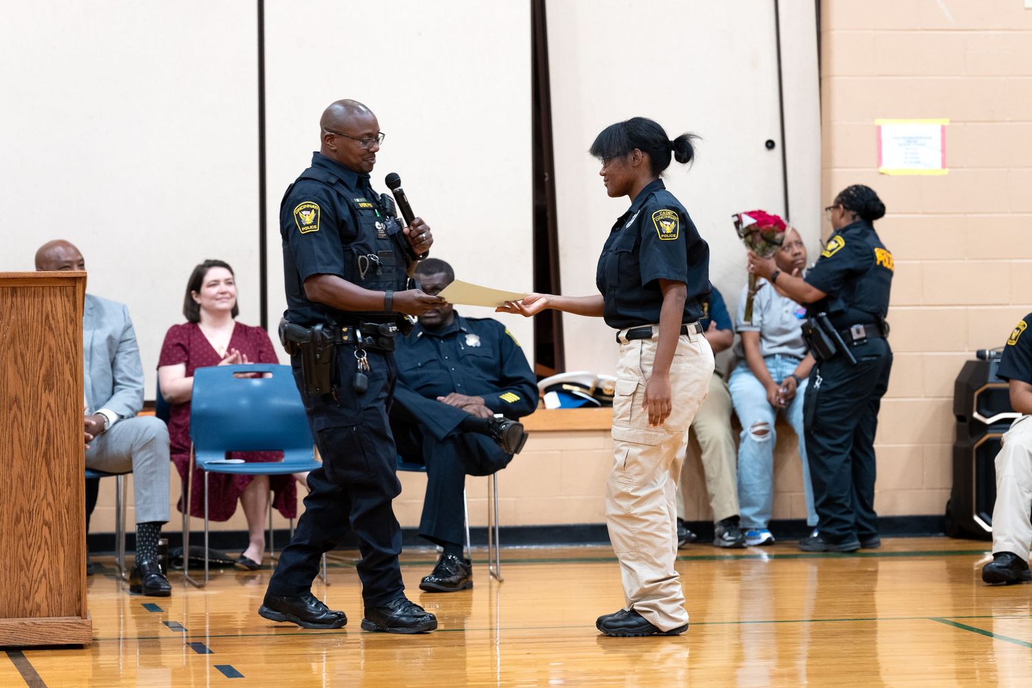 Police officers in uniform exchange items during a ceremony in a school gymnasium with seated audience members.