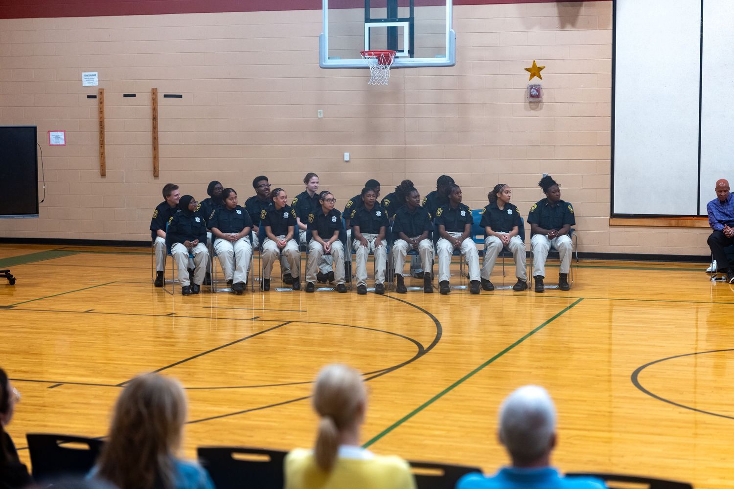 Youth basketball team seated in a row on a gym court during a school assembly or sports event.