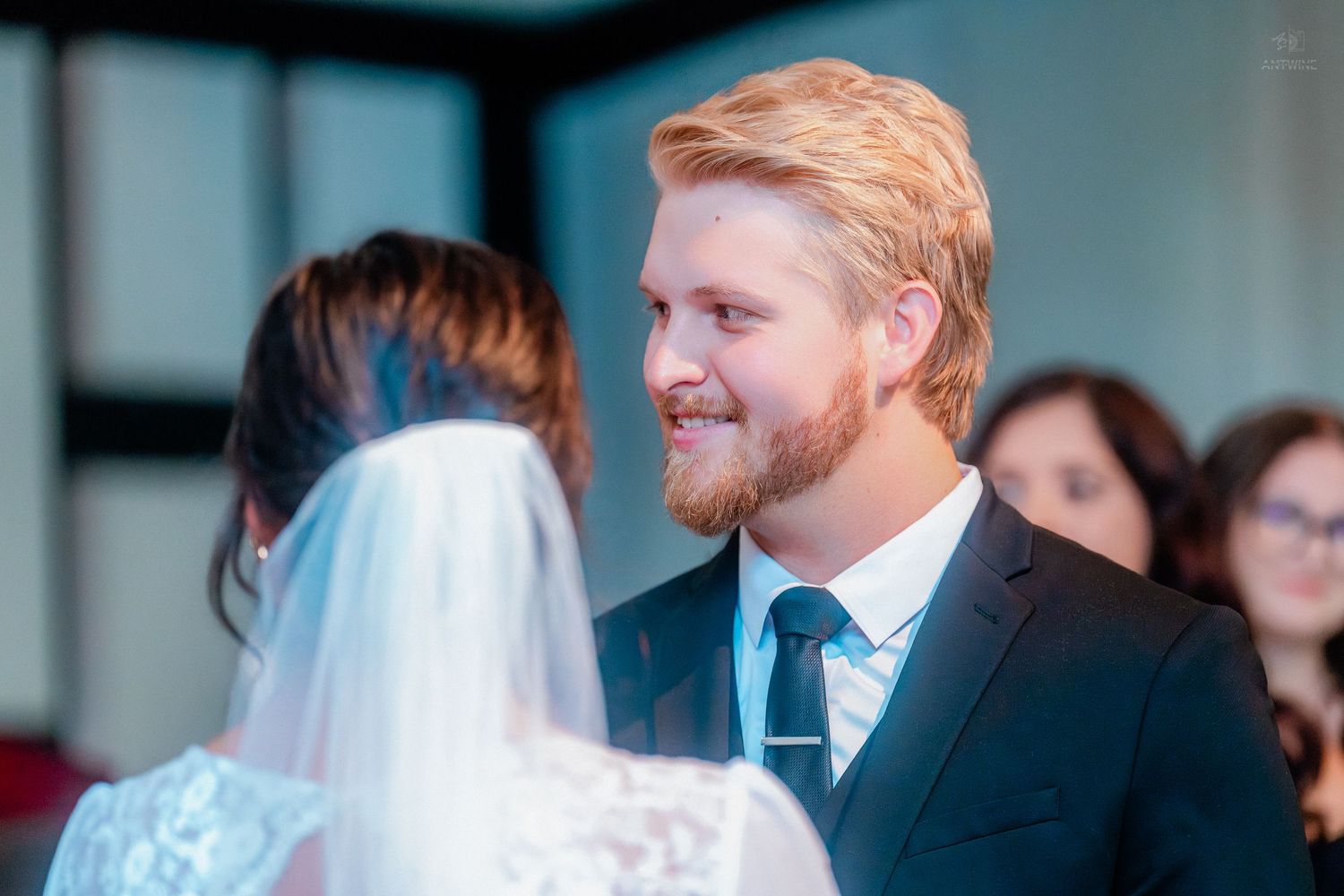 Smiling groom in dark suit and teal tie facing bride in white lace dress and veil at wedding ceremony.