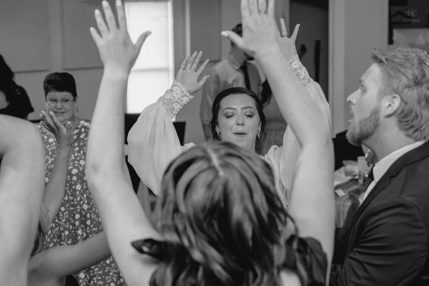 Black and white photo of wedding guests dancing with hands raised in celebration at a lively reception.