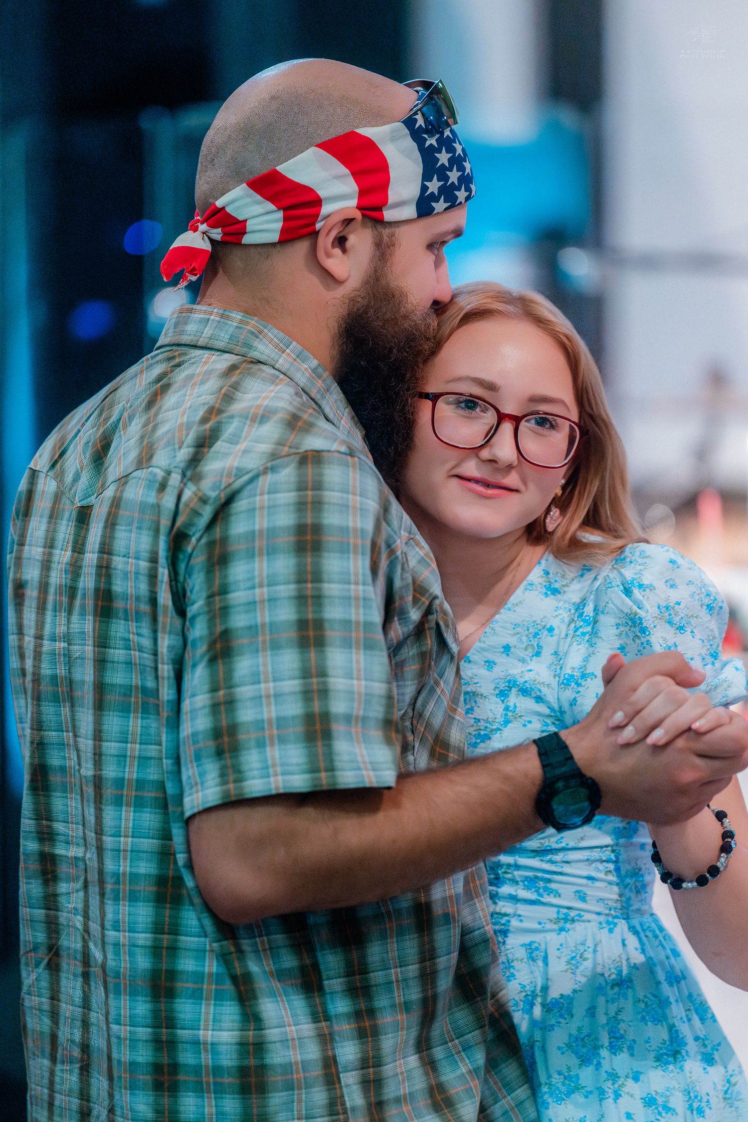 Couple dancing together outdoors, man wearing American flag bandana and plaid shirt, woman in blue floral dress with glasses.