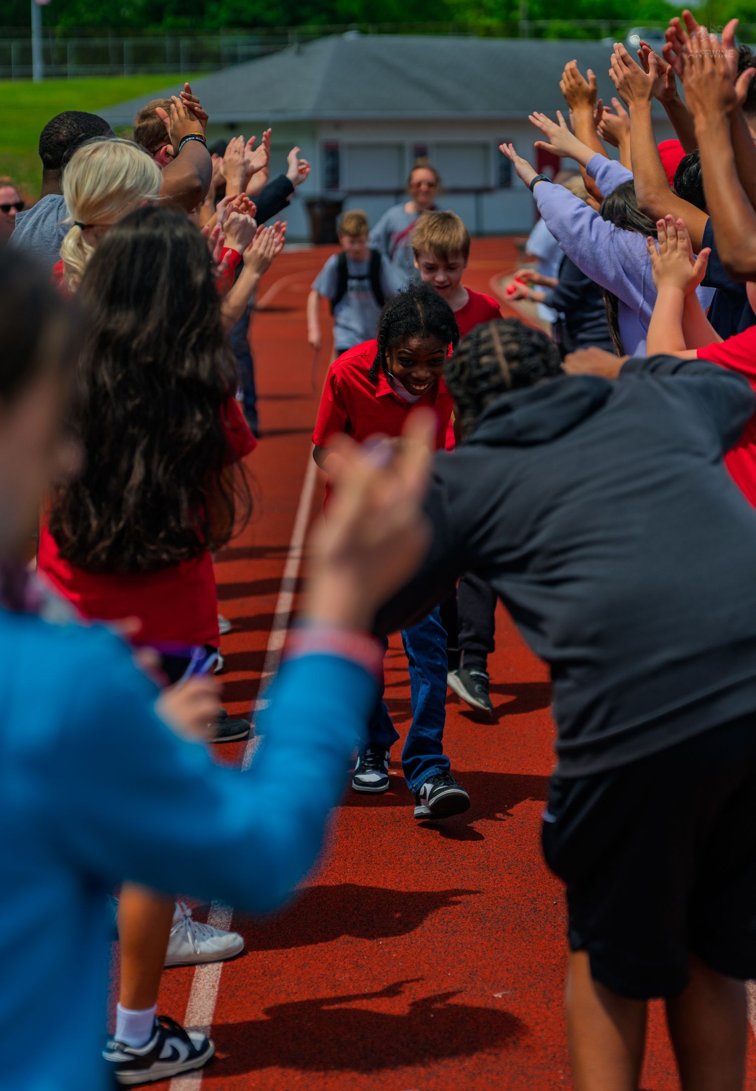 Children running through a celebratory tunnel of raised hands on a red track during a school event.