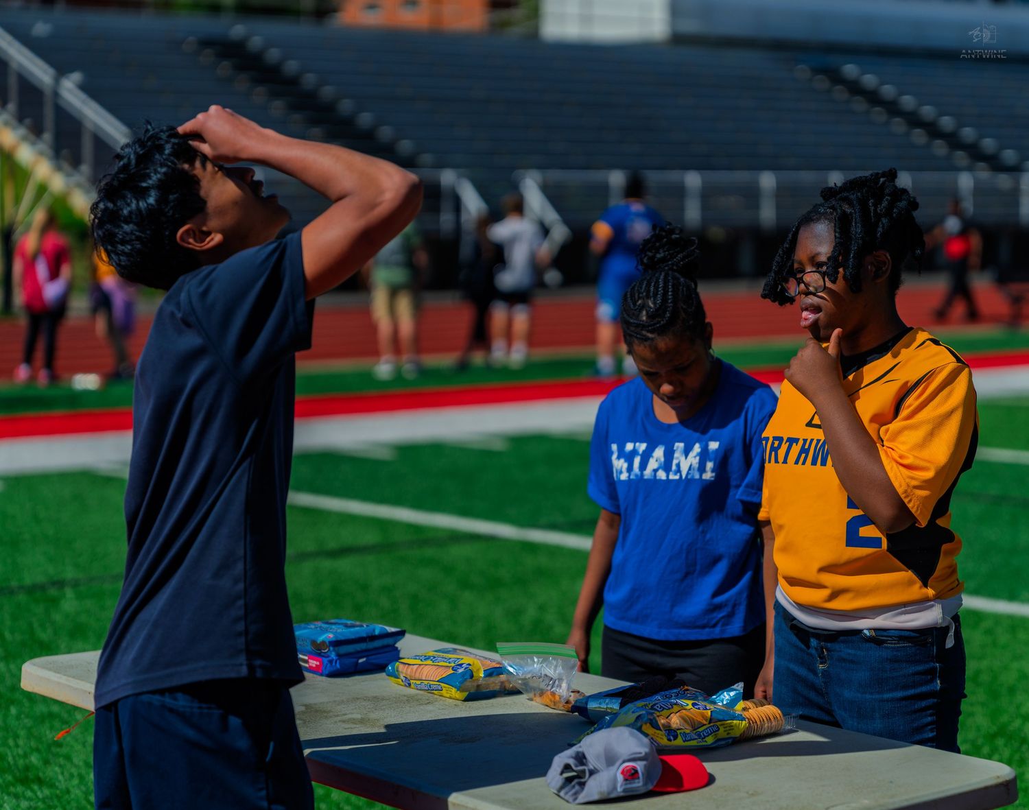 Students gather around a picnic table at a stadium field day event, with one drinking water and others looking on.
