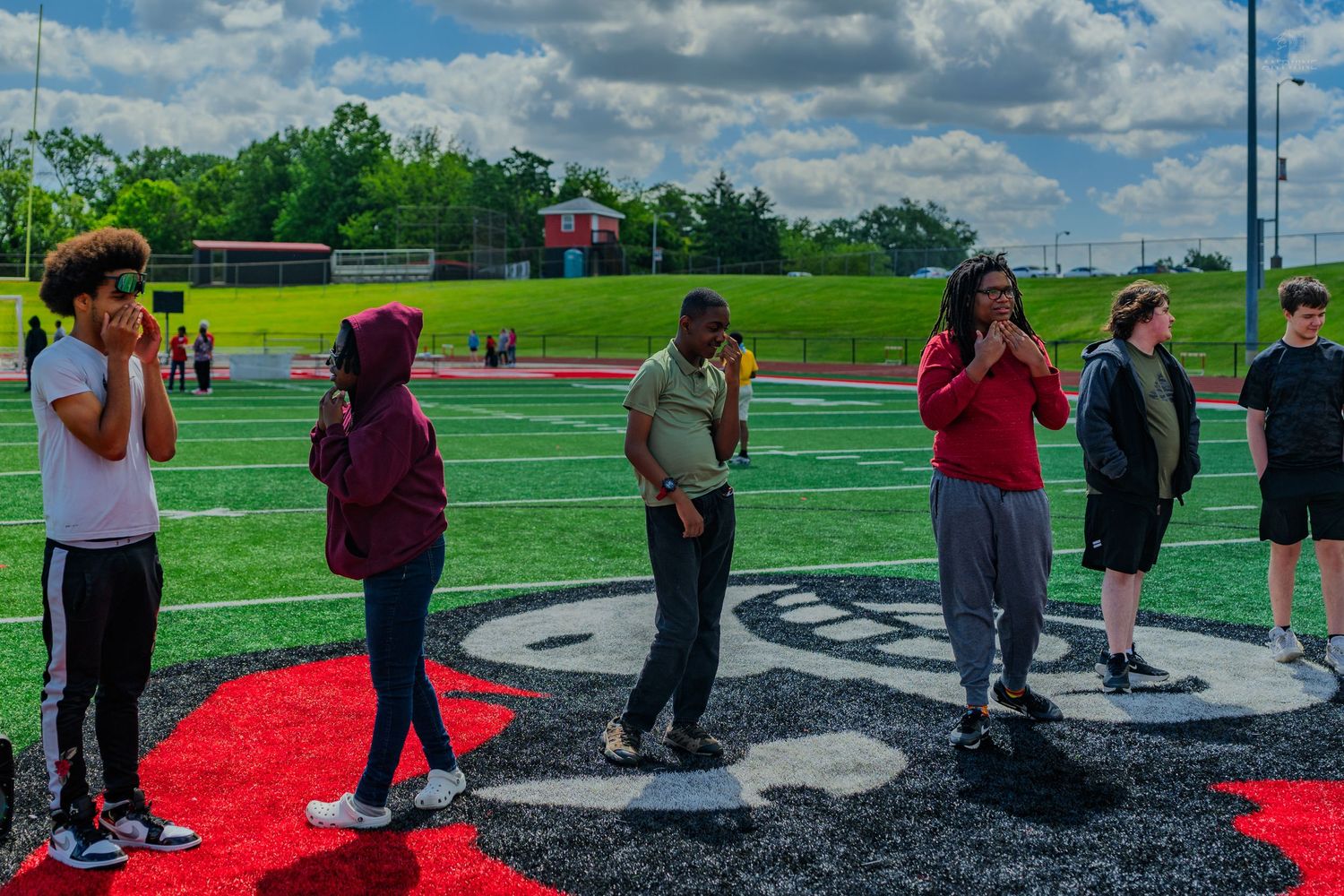 Students stand on a red and black football field logo during an outdoor school activity on a cloudy day.