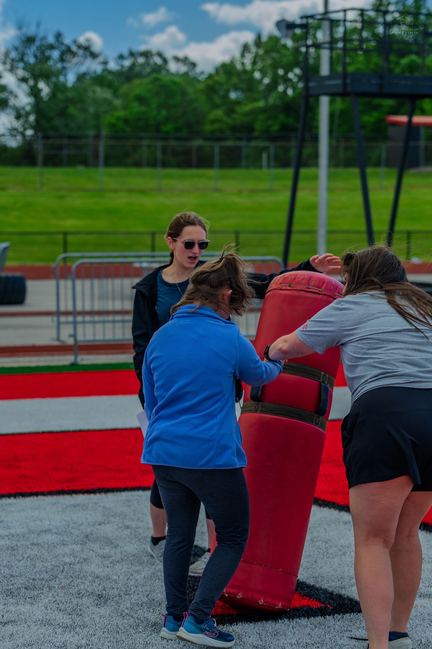 Three women working together to lift a large red punching bag outdoors at a sports facility.