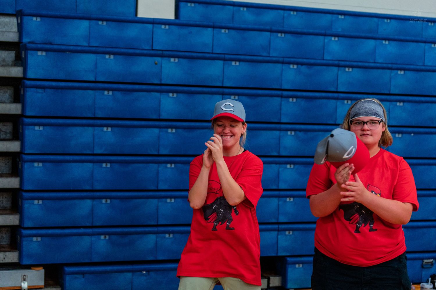 Two women in red shirts clapping and cheering in front of blue bleachers at a sports event.