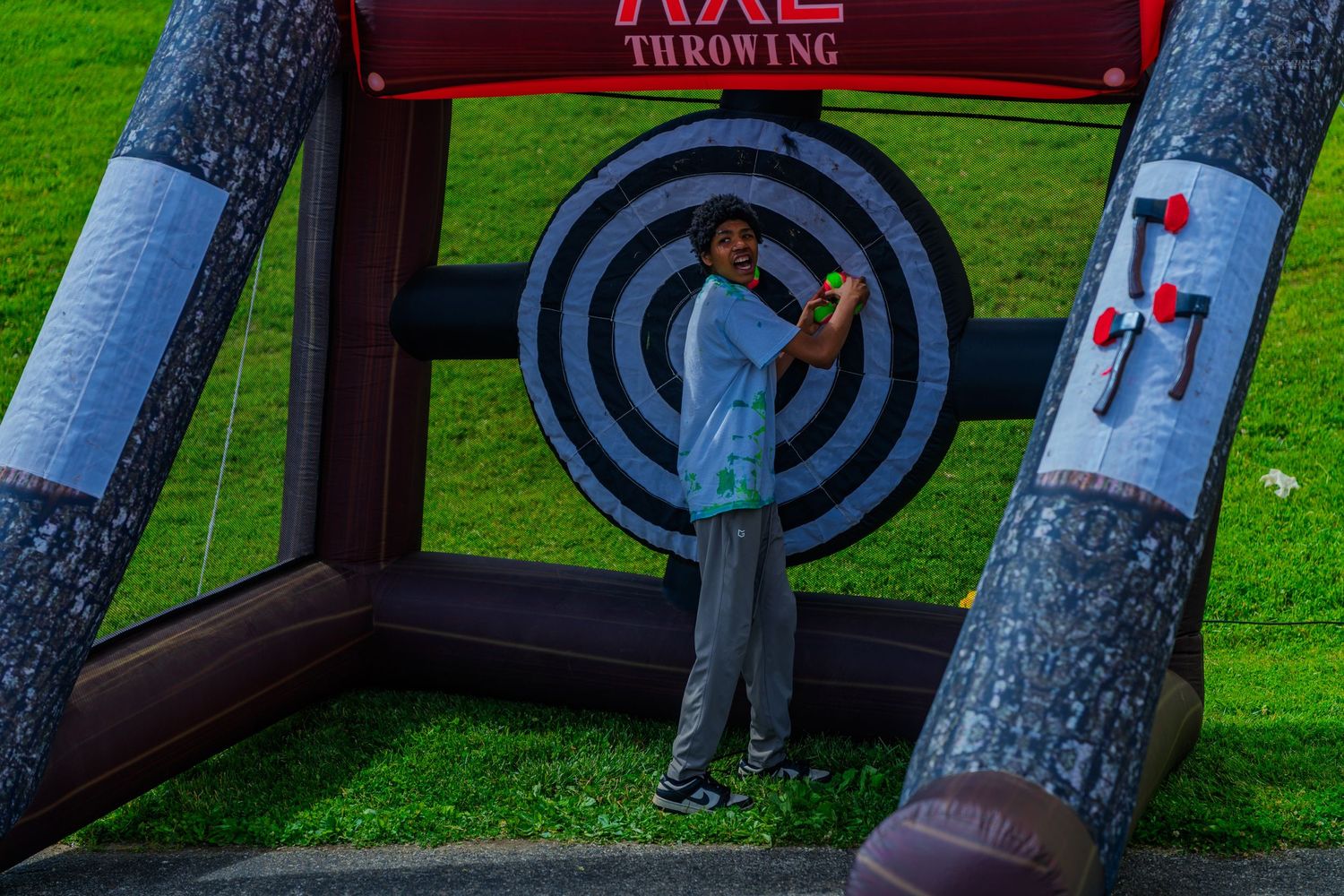 Child poses at inflatable axe throwing game attraction on green grass at outdoor carnival event.