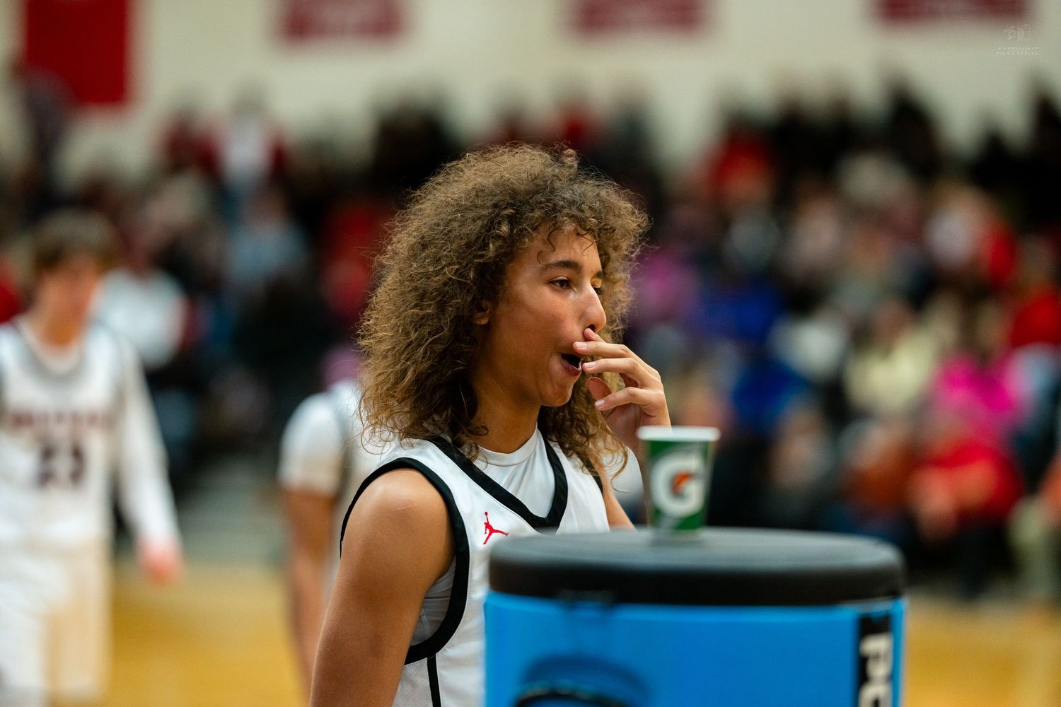 Curly-haired basketball player drinks from cup during break in high school game.