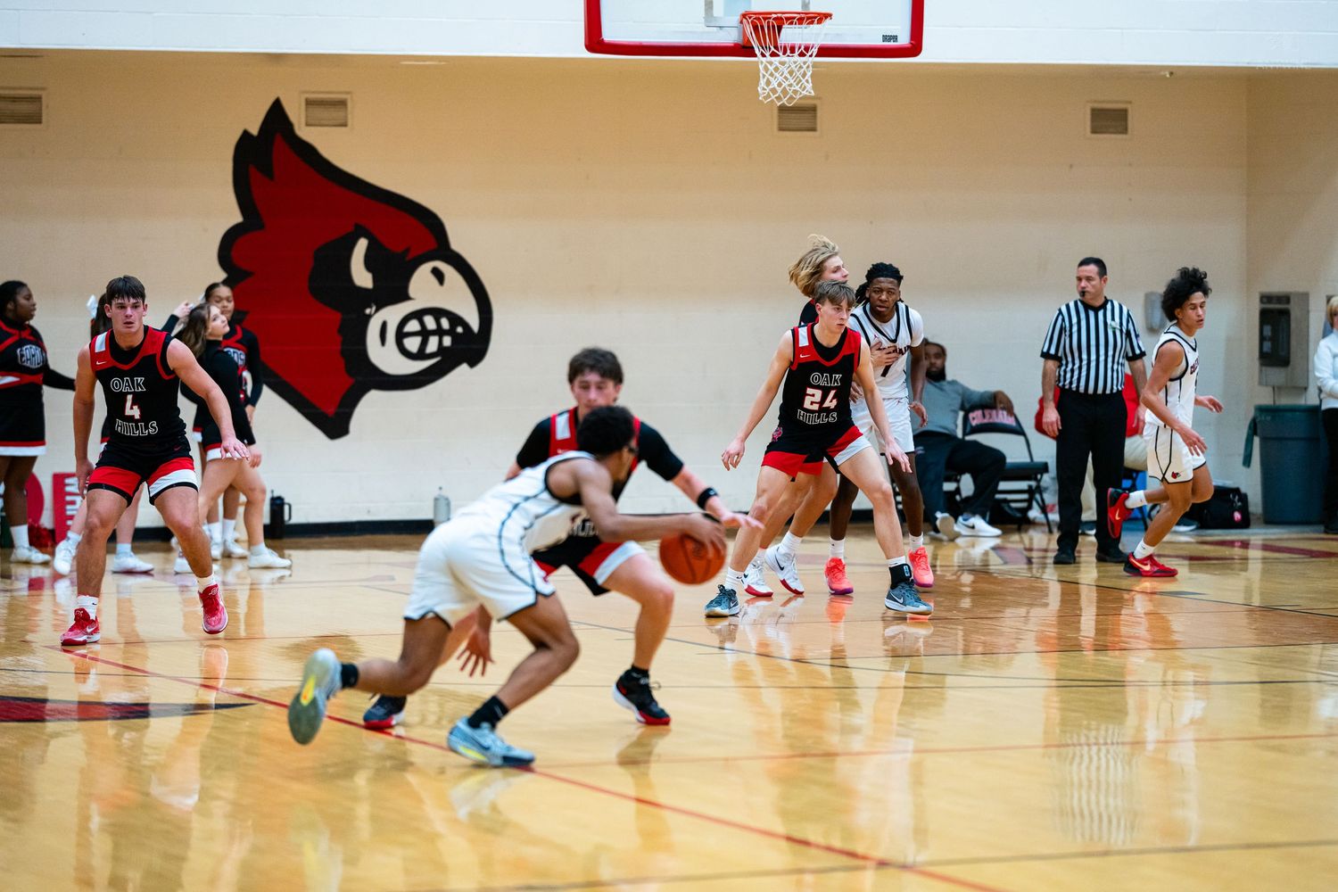 Female basketball players in red and white uniforms compete on an indoor court with a Cardinal mascot logo on the wall.