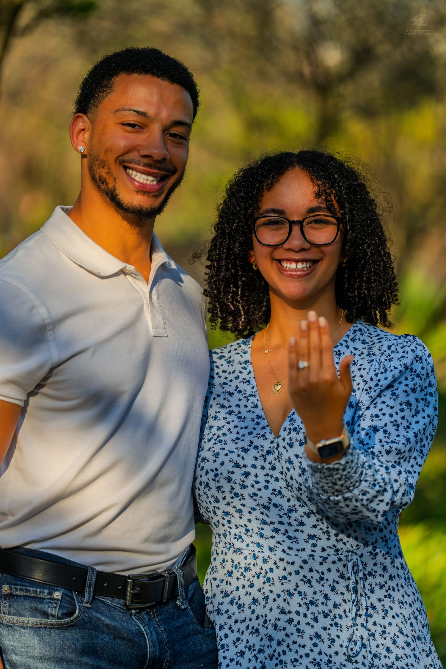 Happy couple showing off engagement ring outdoors, woman in blue floral dress displaying diamond ring while smiling.