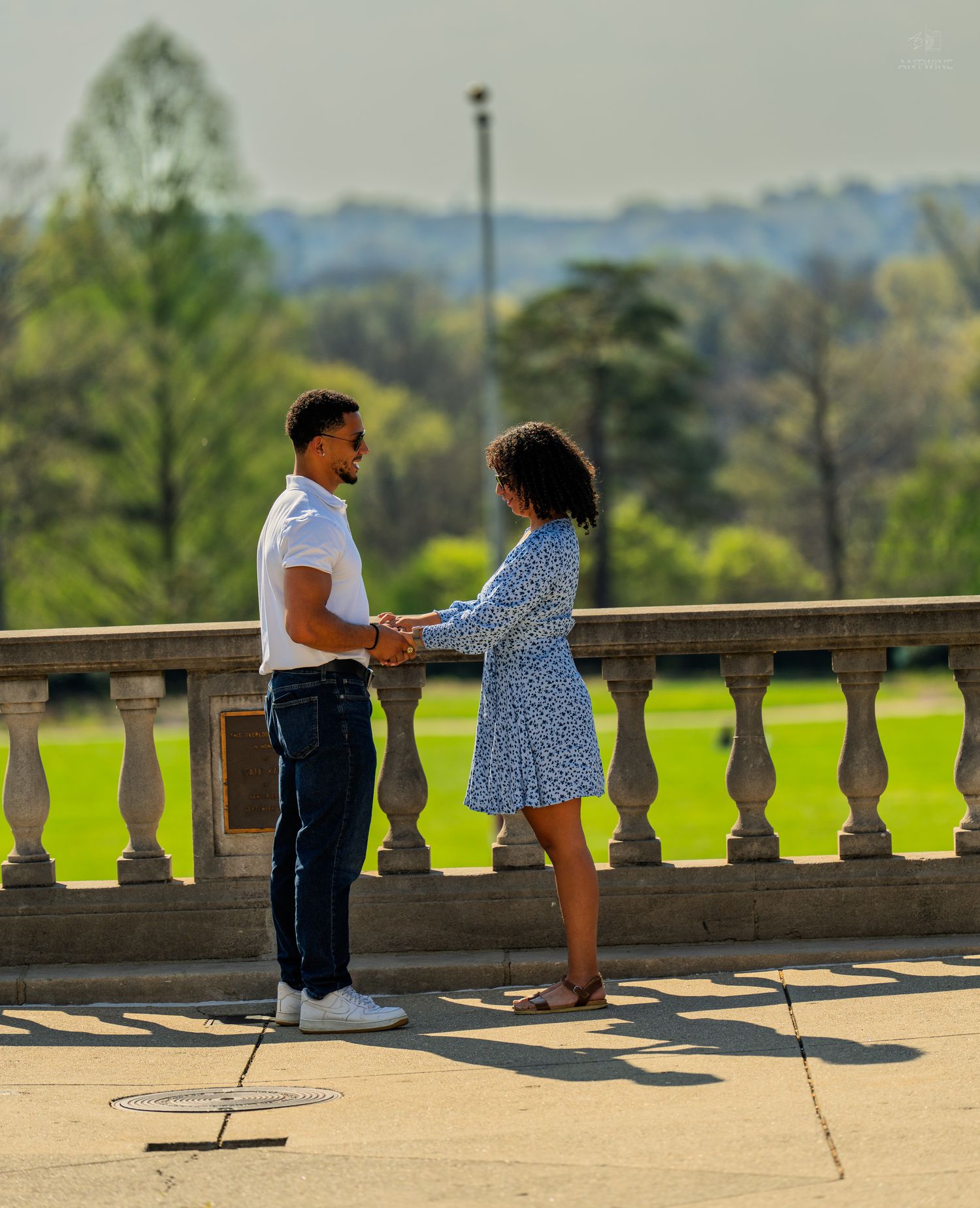 Couple holding hands on a sunny terrace with stone balustrade and green park backdrop.