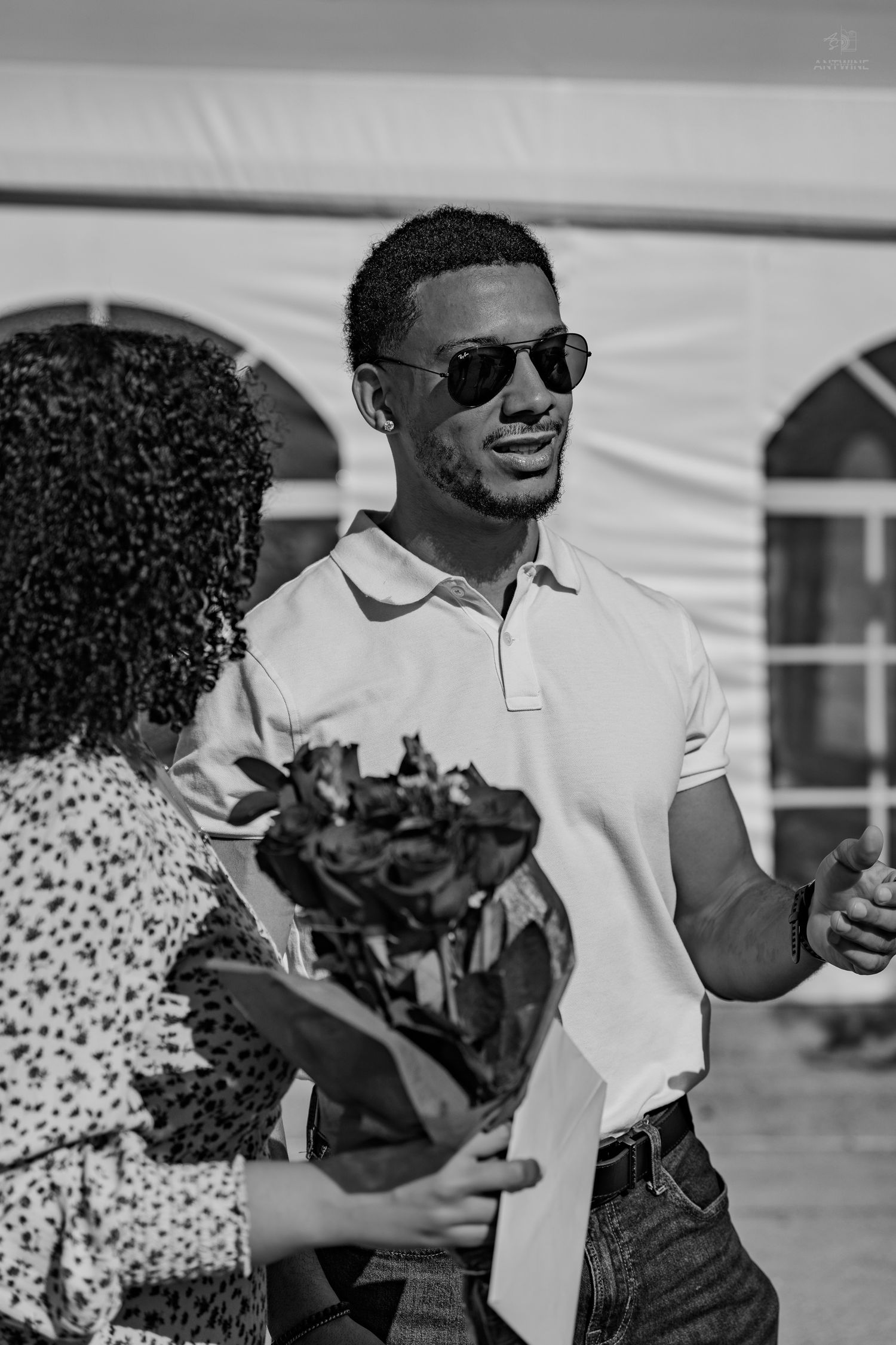 A smiling man in sunglasses and white polo receives a bouquet of flowers outdoors in black and white.