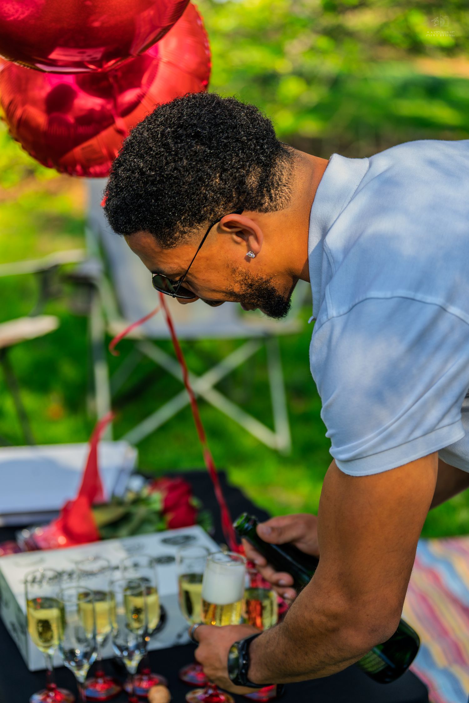 Man pouring champagne at a decorated outdoor table with red heart balloons.