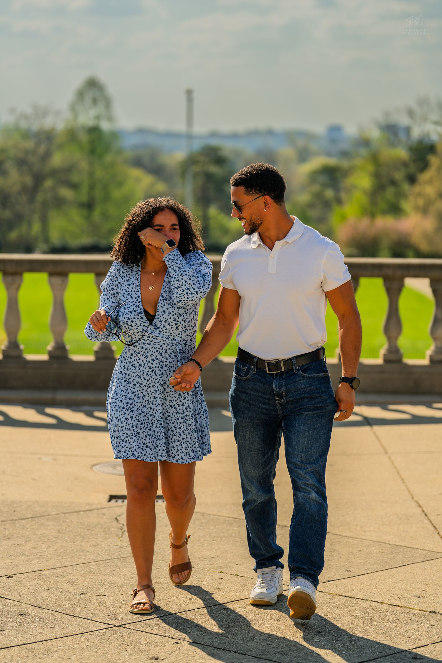 A couple walks hand in hand on a sunlit stone terrace, smiling at each other with park views.