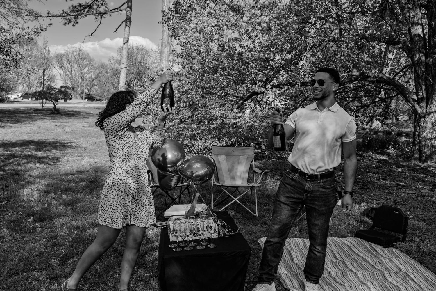 Couple celebrating outdoors in black and white, woman raising bottle while man holds drink in park setting.
