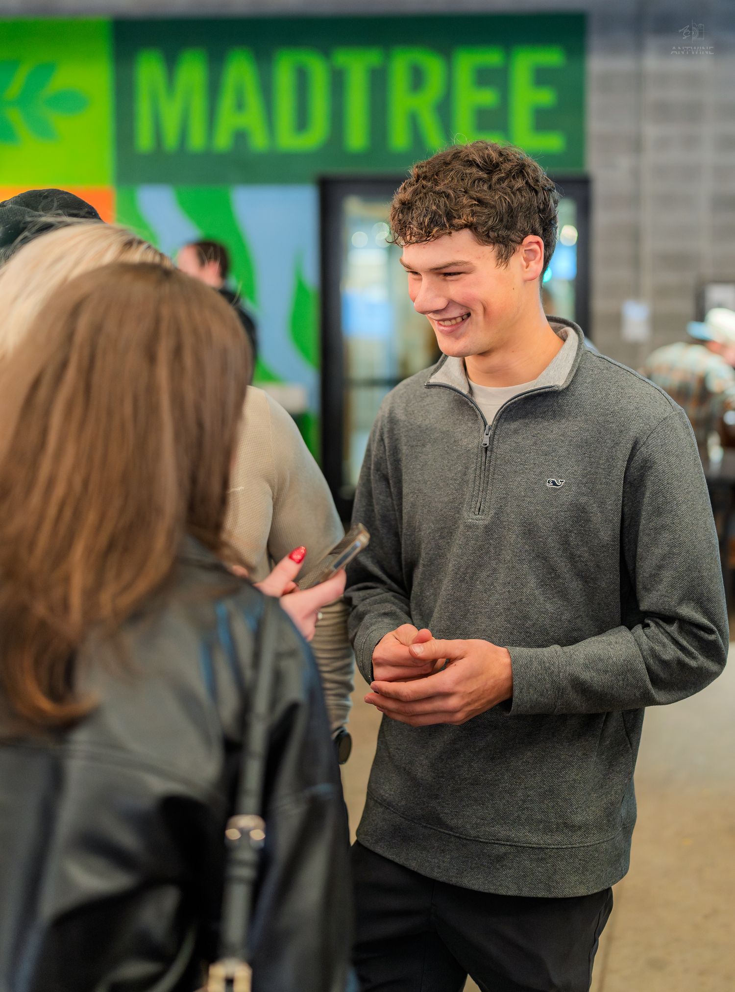 Smiling young man in gray quarter-zip sweater standing outside MadTree Brewing holding a phone.