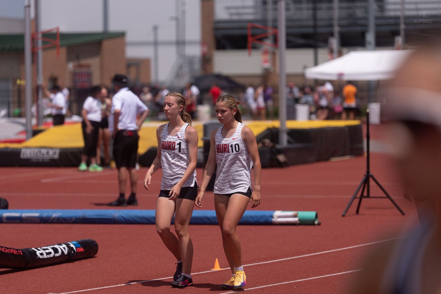 Two female track athletes wearing matching uniforms stand together on a red track at an outdoor state championship meet.