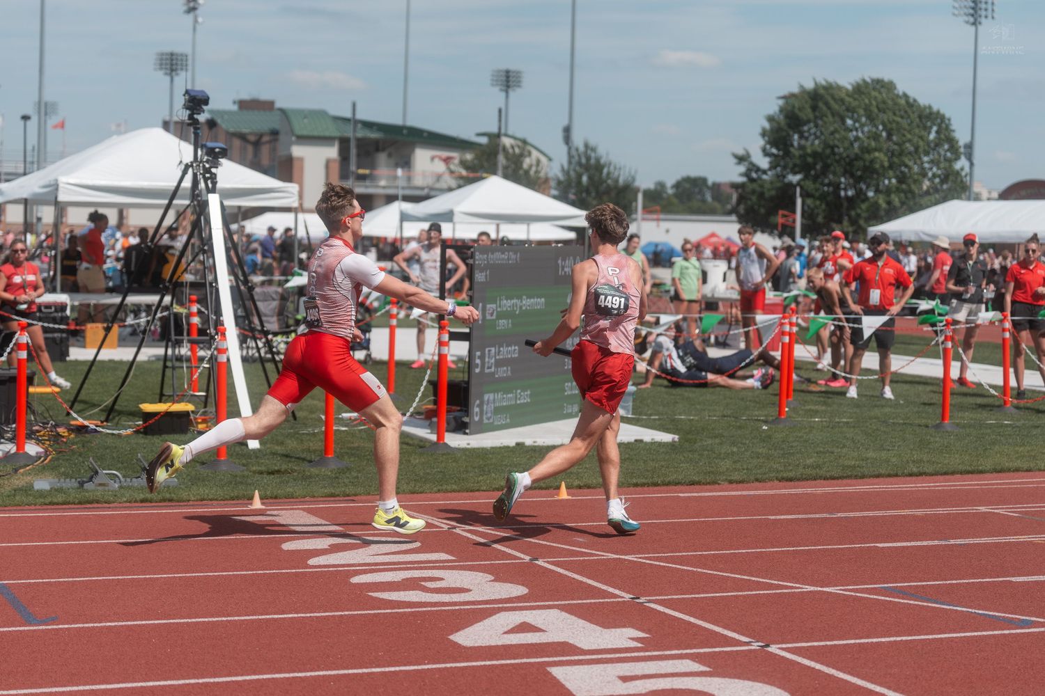 Relay runners exchange a baton near the finish line at a sunny outdoor high school track and field meet.