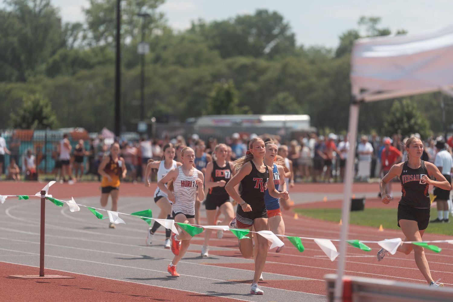Female athletes sprint toward the finish line at a sunny outdoor track and field championship meet.