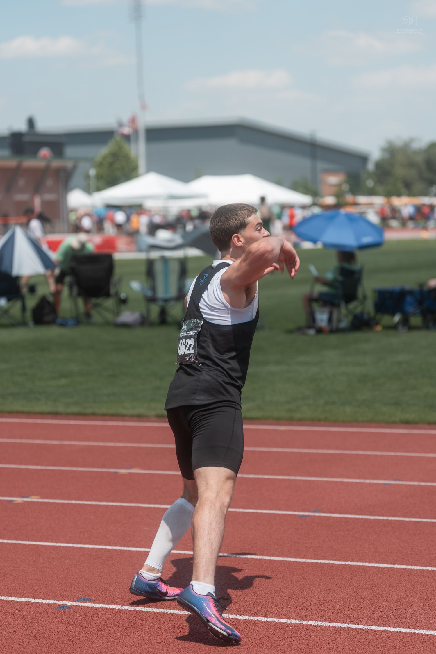 Male athlete in black uniform prepares to throw a shot put on a red track at an outdoor track and field competition.