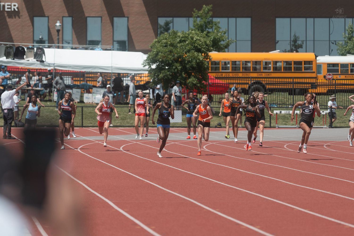Female sprinters racing on red track at outdoor collegiate track and field meet near brick building.