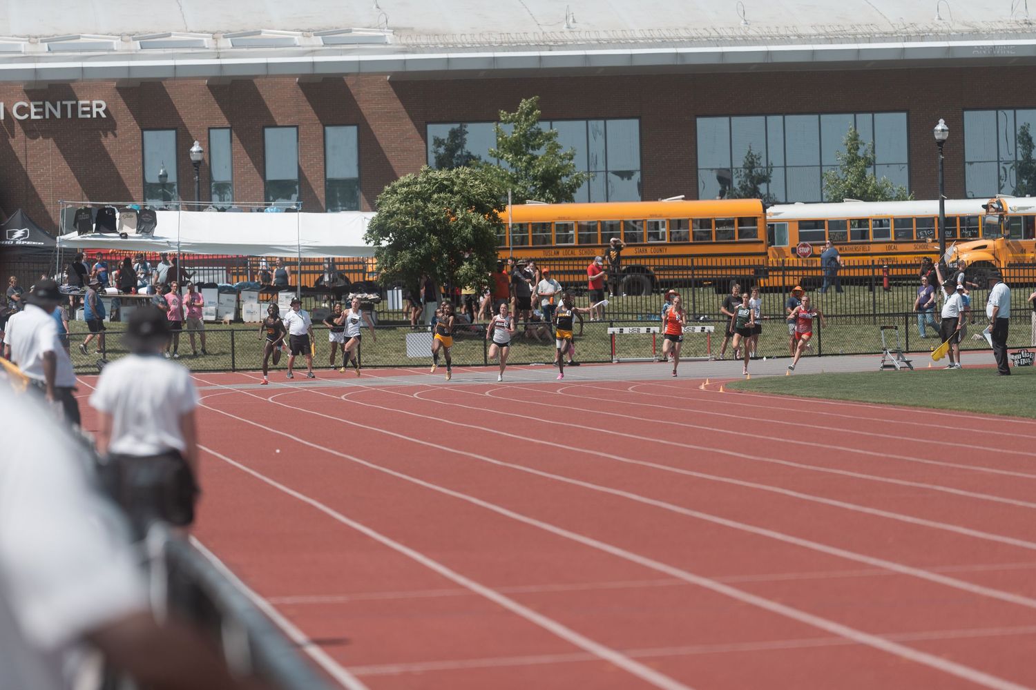 Runners compete on a red track with yellow school buses and a modern sports center in the background.