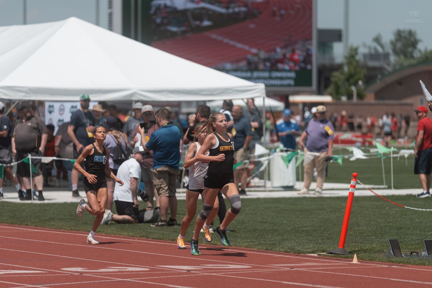 Athletes compete in a track and field race at an outdoor stadium during a sunny day meet.