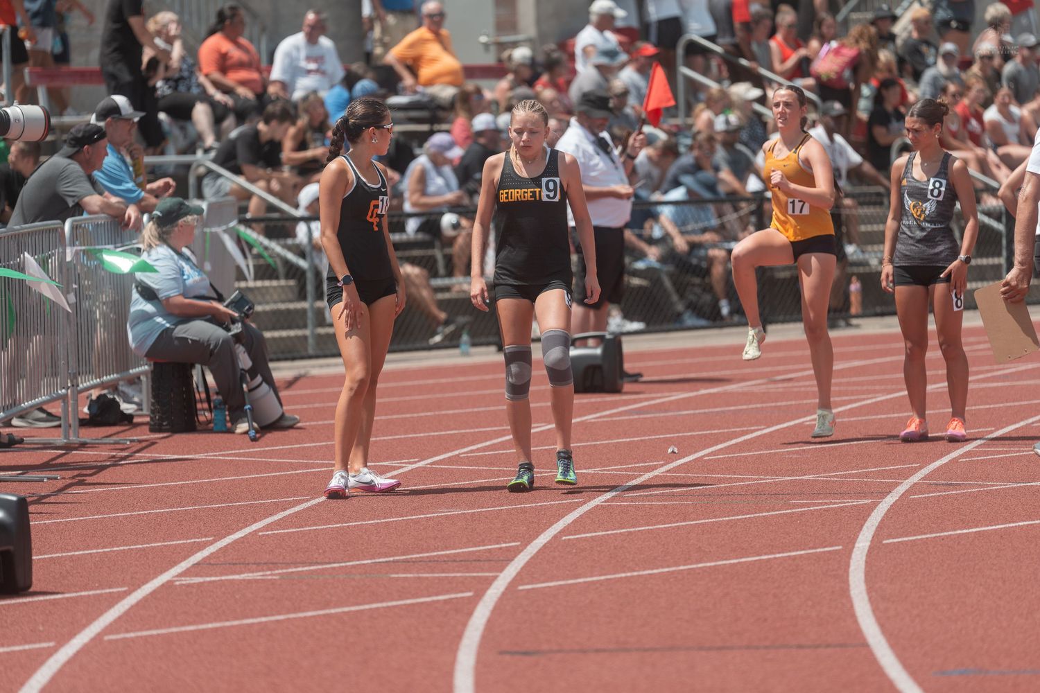 Young female runners prepare on a red track during a high school track and field competition with spectators watching.