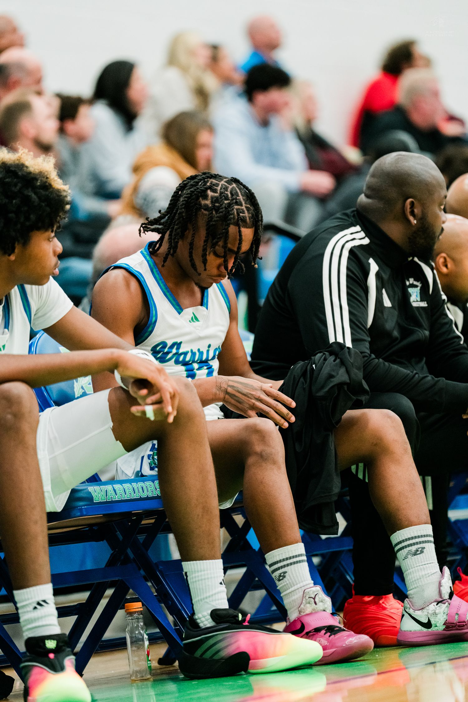 A high school basketball player in white Warriors uniform sits on the bench with coaches during a game.