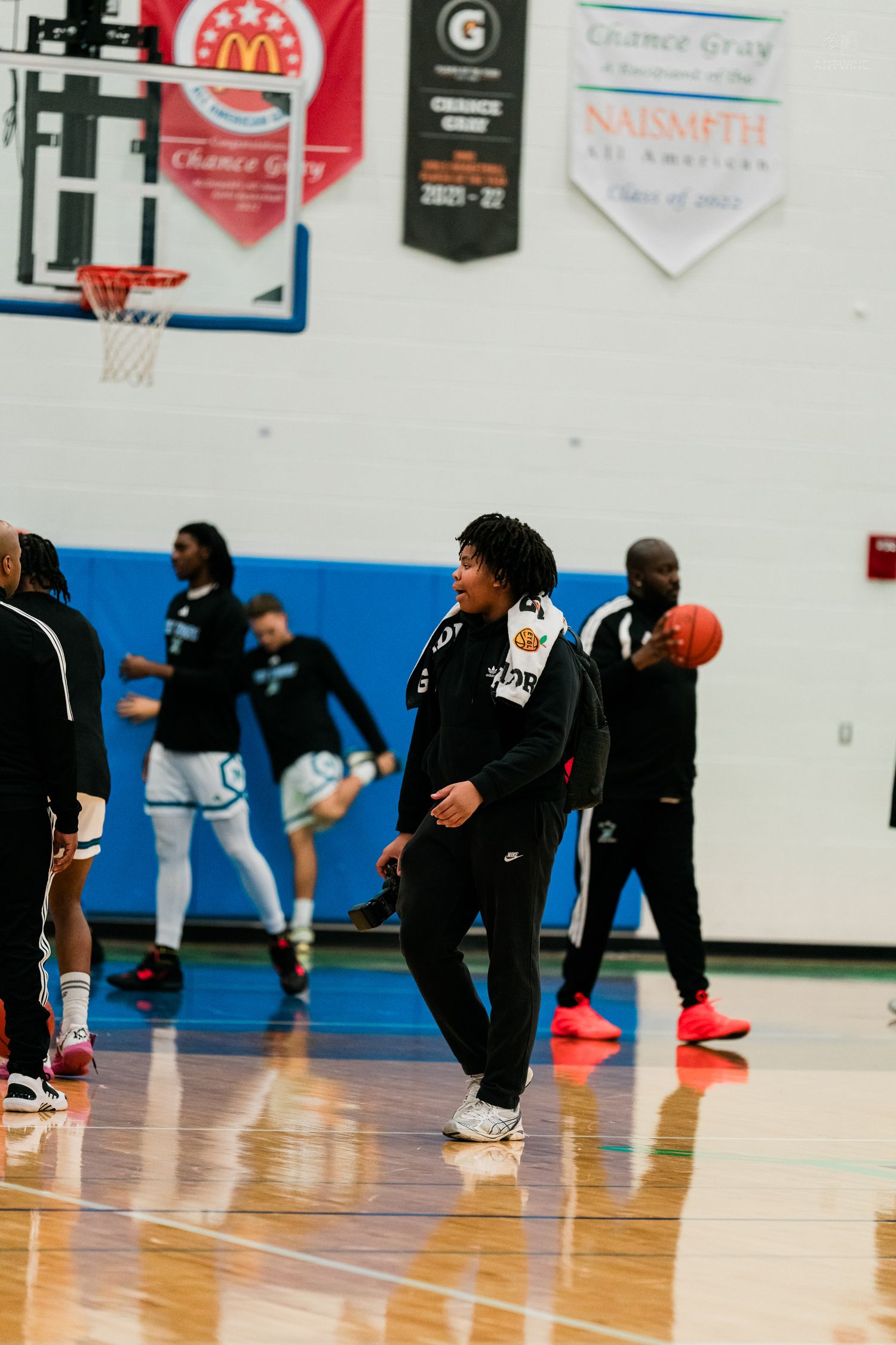 A young basketball player in black warmup gear walks on a court during pre-game warm-ups.